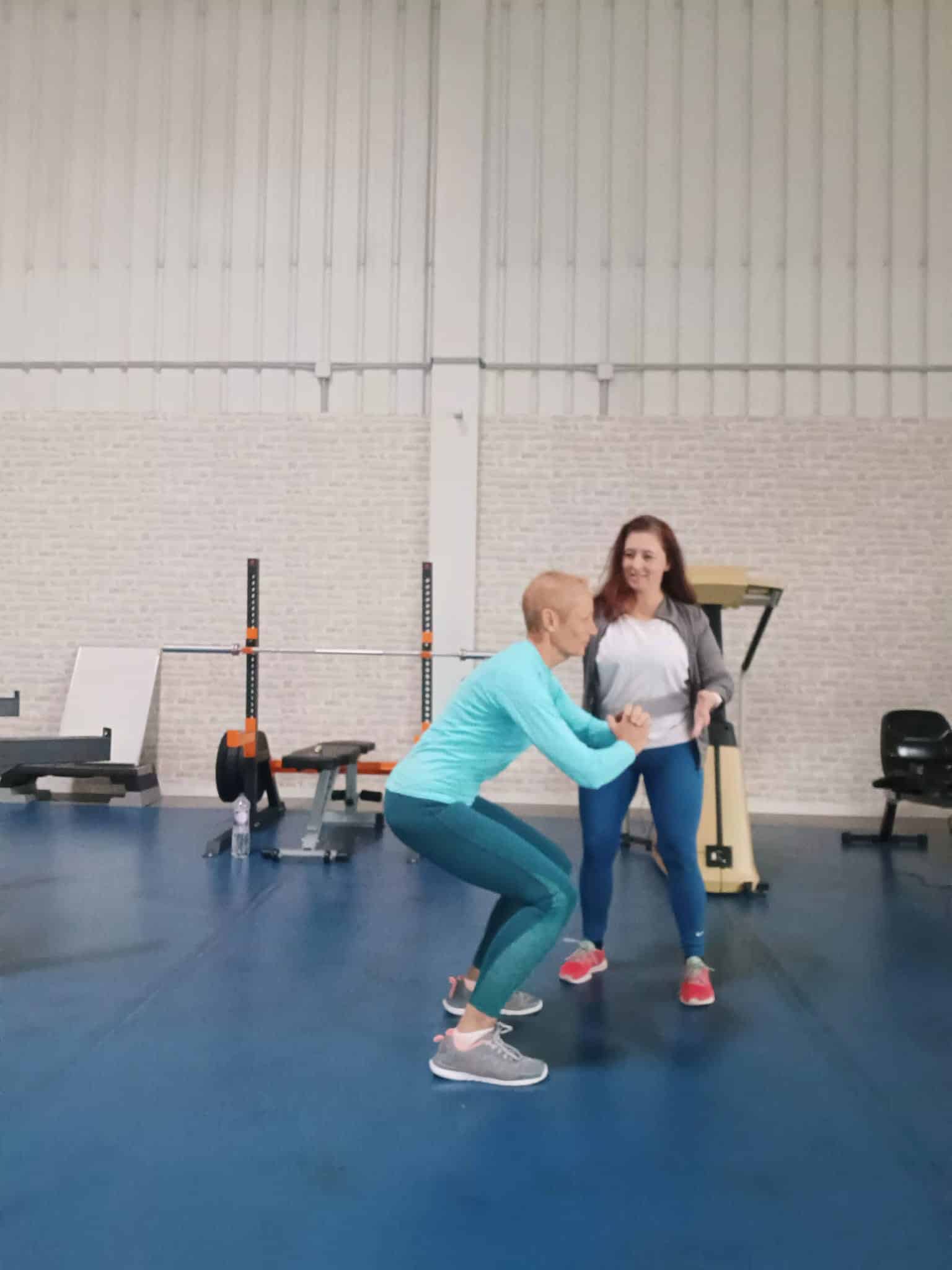 Femme en squat assistée par une coach dans une salle de sport lumineuse avec équipements de musculation.