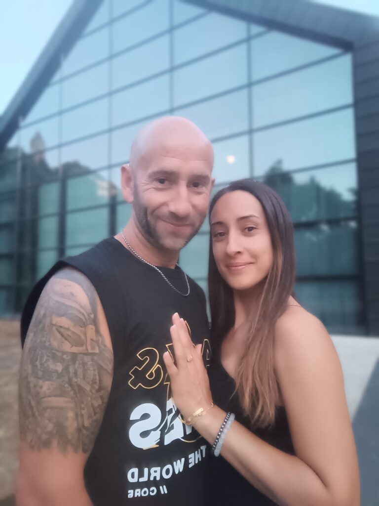 Couple souriant devant un bâtiment vitré, homme tatoué et femme aux bijoux dorés.