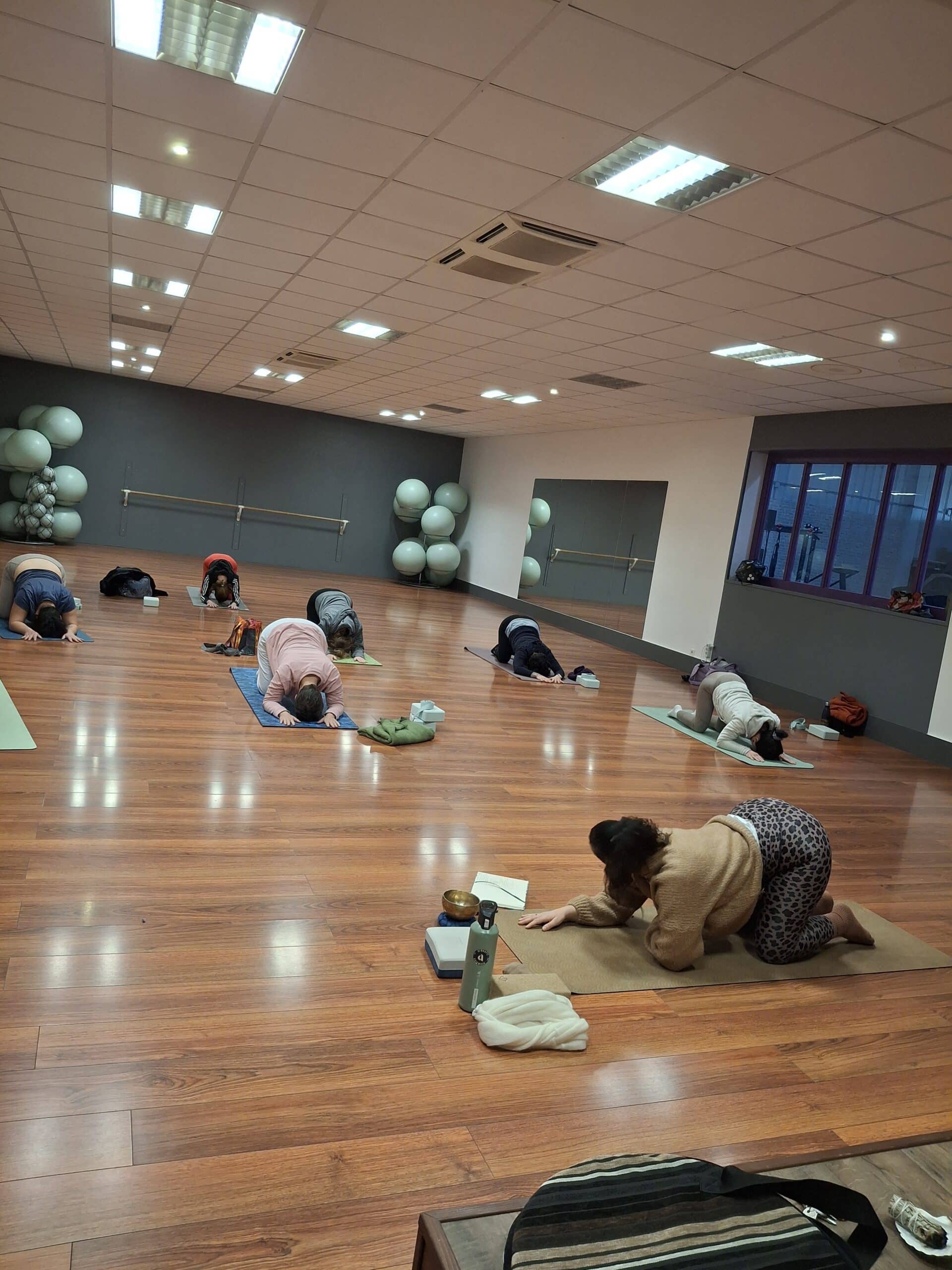 Personnes en posture de l'enfant (Balasana) sur tapis dans un studio de yoga avec parquet et ballons de gym.