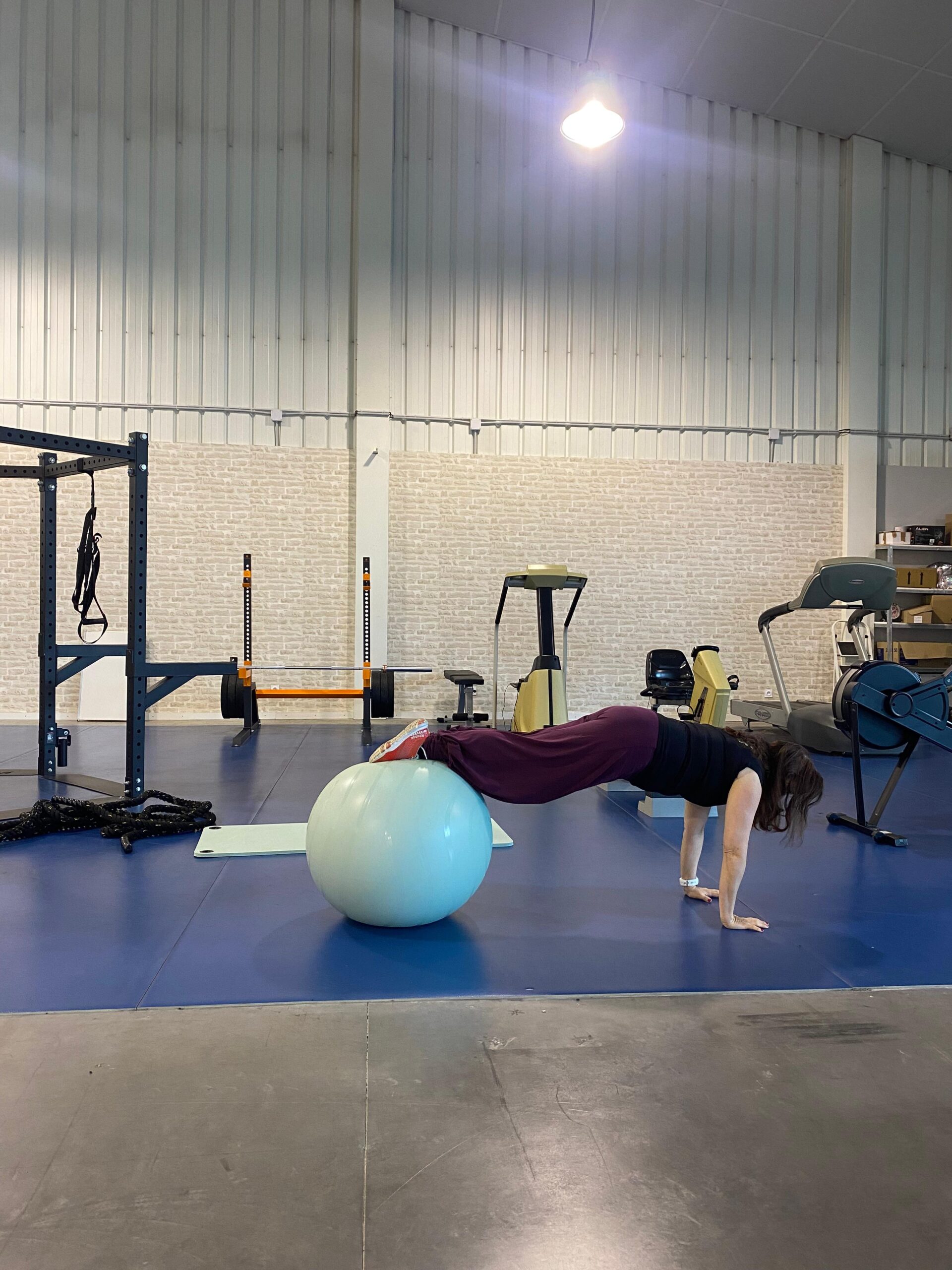 Femme en gainage sur ballon de gym bleu, mains au sol, dans une salle de sport spacieuse.