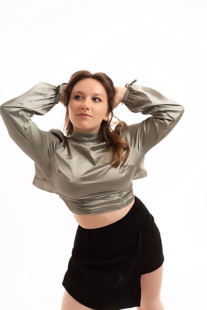 Femme en haut satiné vert sauge et jupe noire posant mains derrière la tête sur fond blanc.