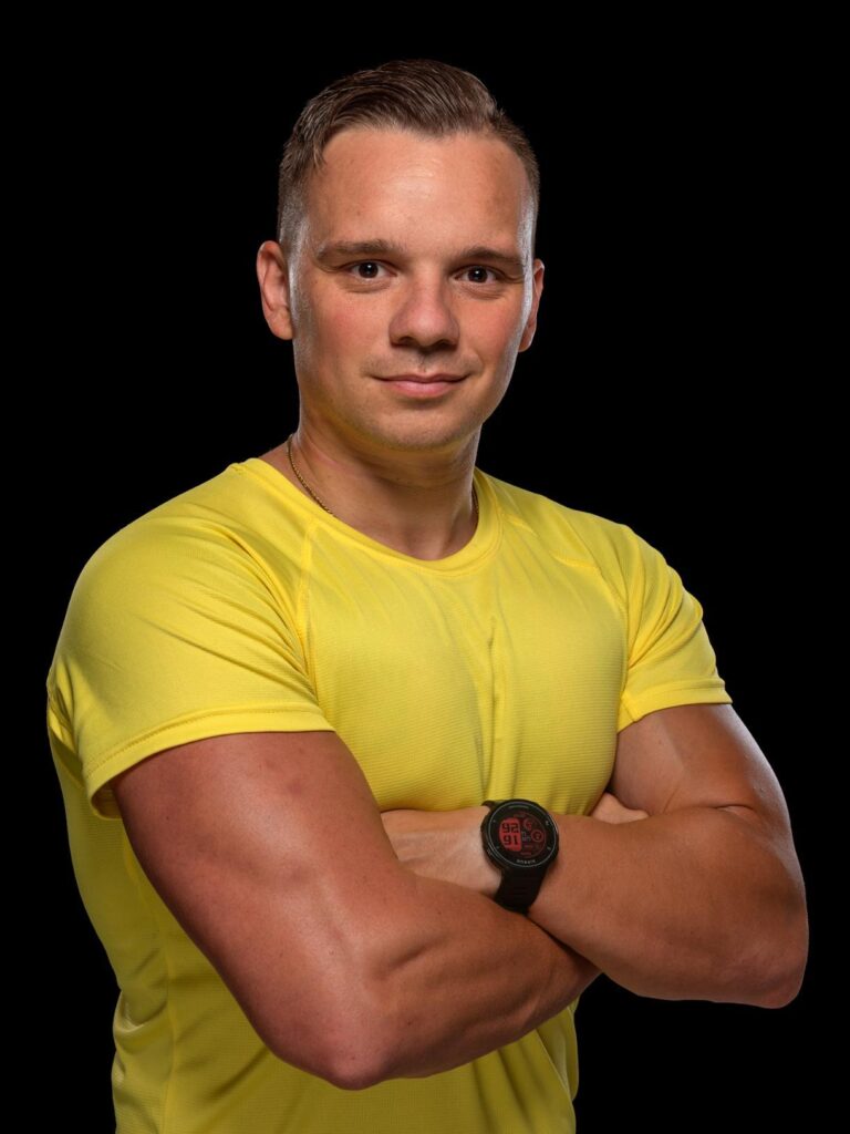 Homme athlétique en t-shirt jaune, bras croisés, portant une montre de sport sur fond noir.