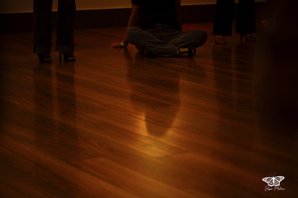 Reflets sur parquet lustré, personne assise en tailleur et jambes d'invités dans une ambiance tamisée.