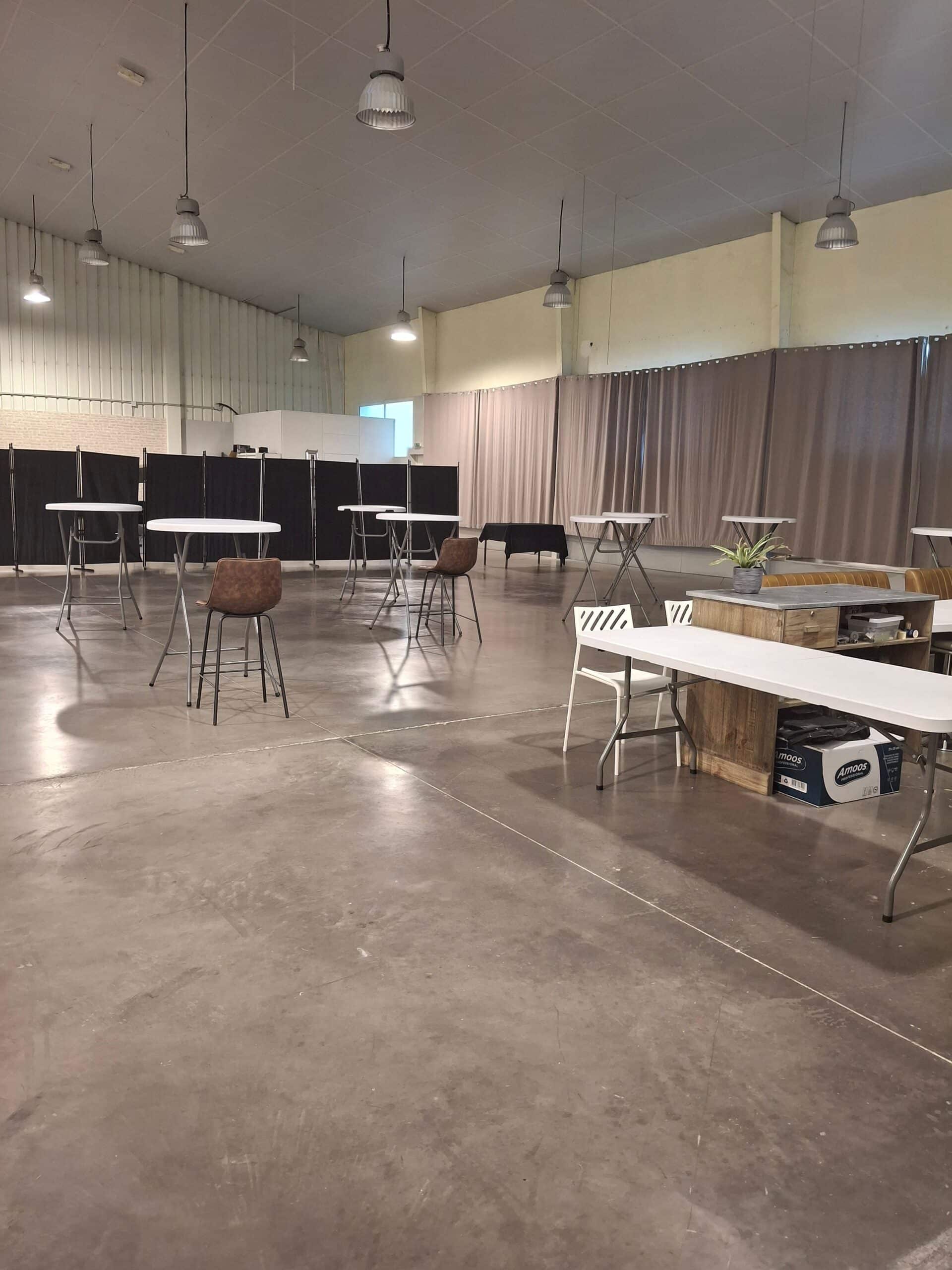 Grande salle événementielle industrielle aménagée avec tables hautes, tabourets et cloisons de séparation.