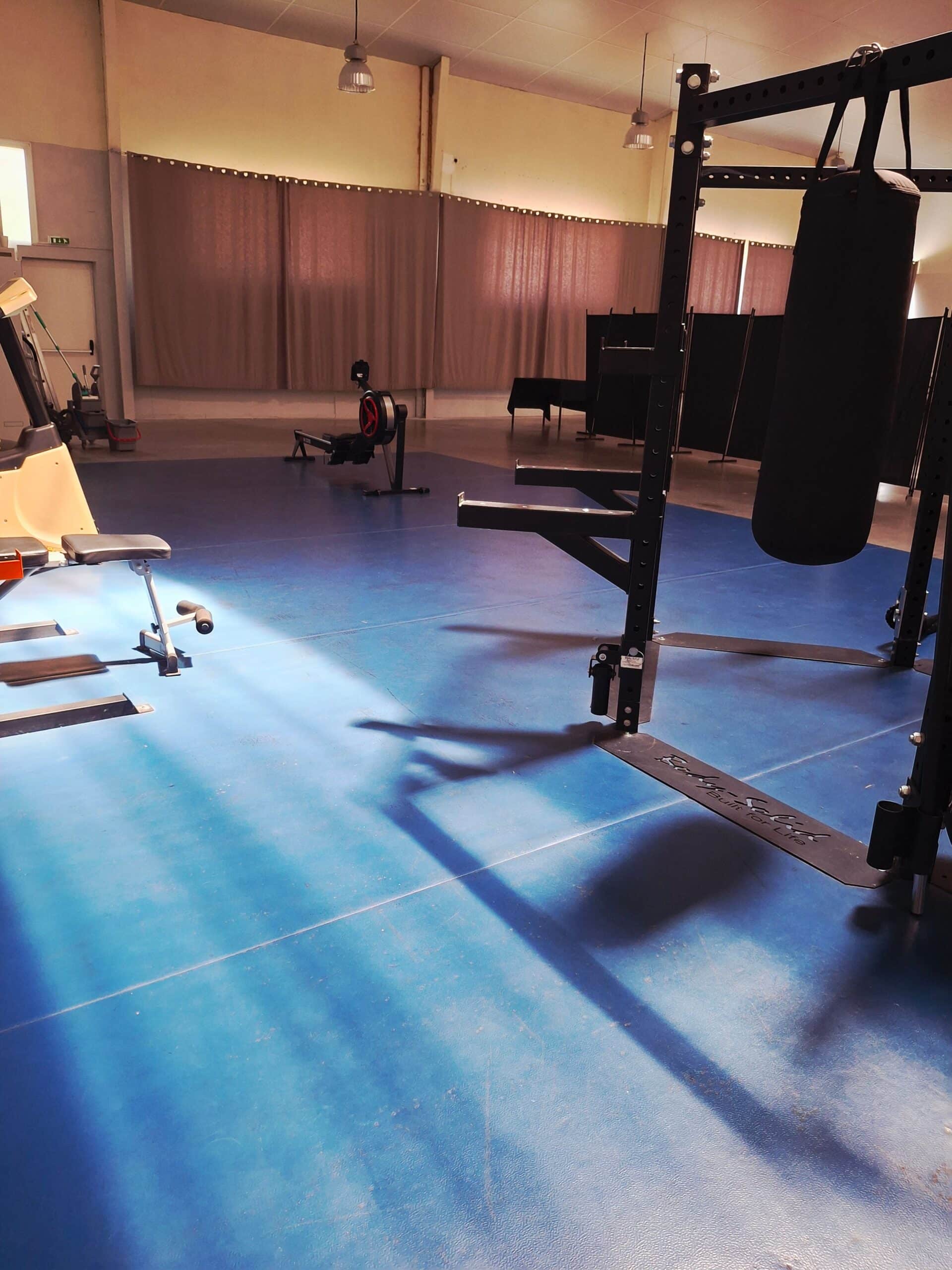 Salle de sport au sol bleu avec rack Body-Solid, sac de frappe et rameur.
