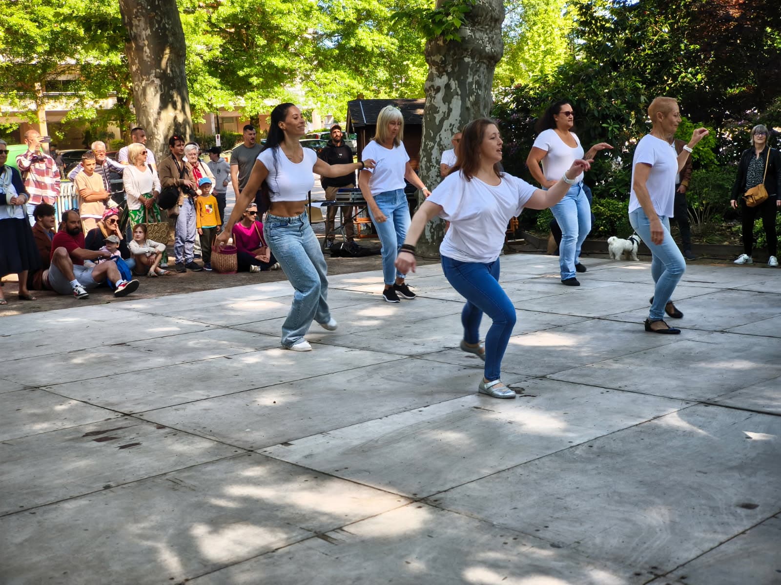 Femmes en blanc et jeans dansant énergiquement en plein air devant un public dans un parc ombragé.