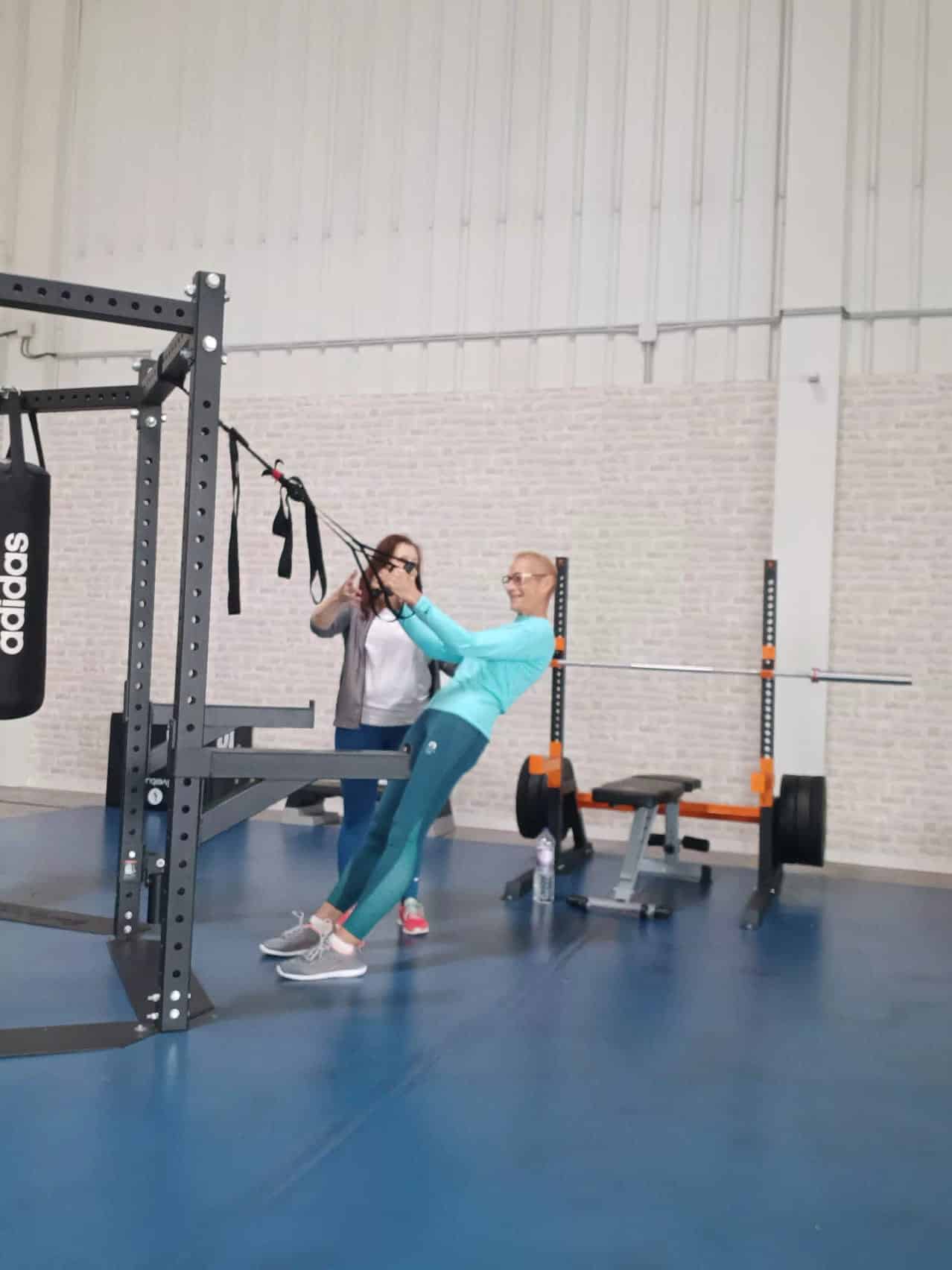 Femme pratiquant le TRX avec une coach en salle de sport, sac Adidas visible.